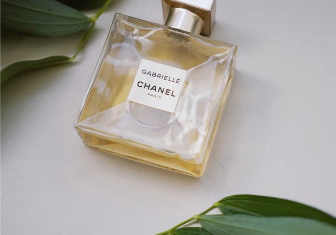 Gabrielle Essence perfume bottle laying flat on a white surface, surrounded by leafy, green plants